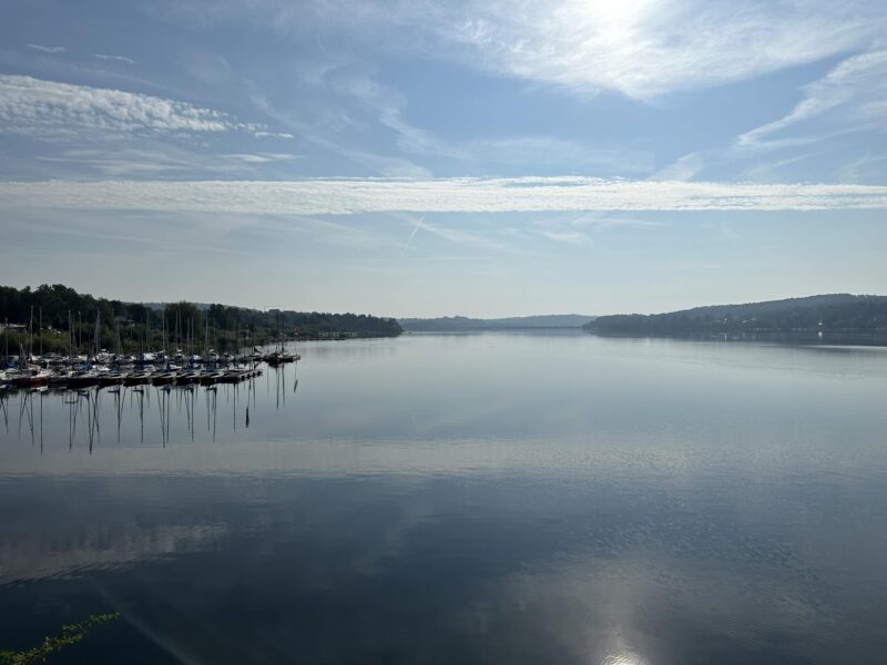 Leichter Dunst über dem Möhnesee am Morgen, Panoramaansicht von einer Fußgängerbrücke fotografiert. Links eine Reihe von Segelboote an zwei Stegen.